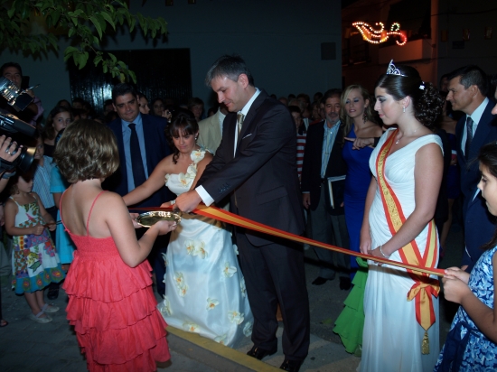 Emi, con vestido blanco de flores, ayuda al alcalde a cortar la cinta inaugural. 